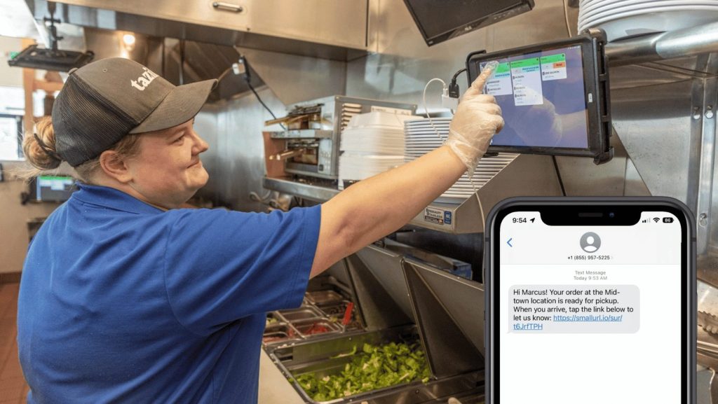 A woman working in a quick-service restaurant's kitchen is tapping an order on a kitchen display system screen and a hone screen showing a notification that the order is ready for pick-up. 